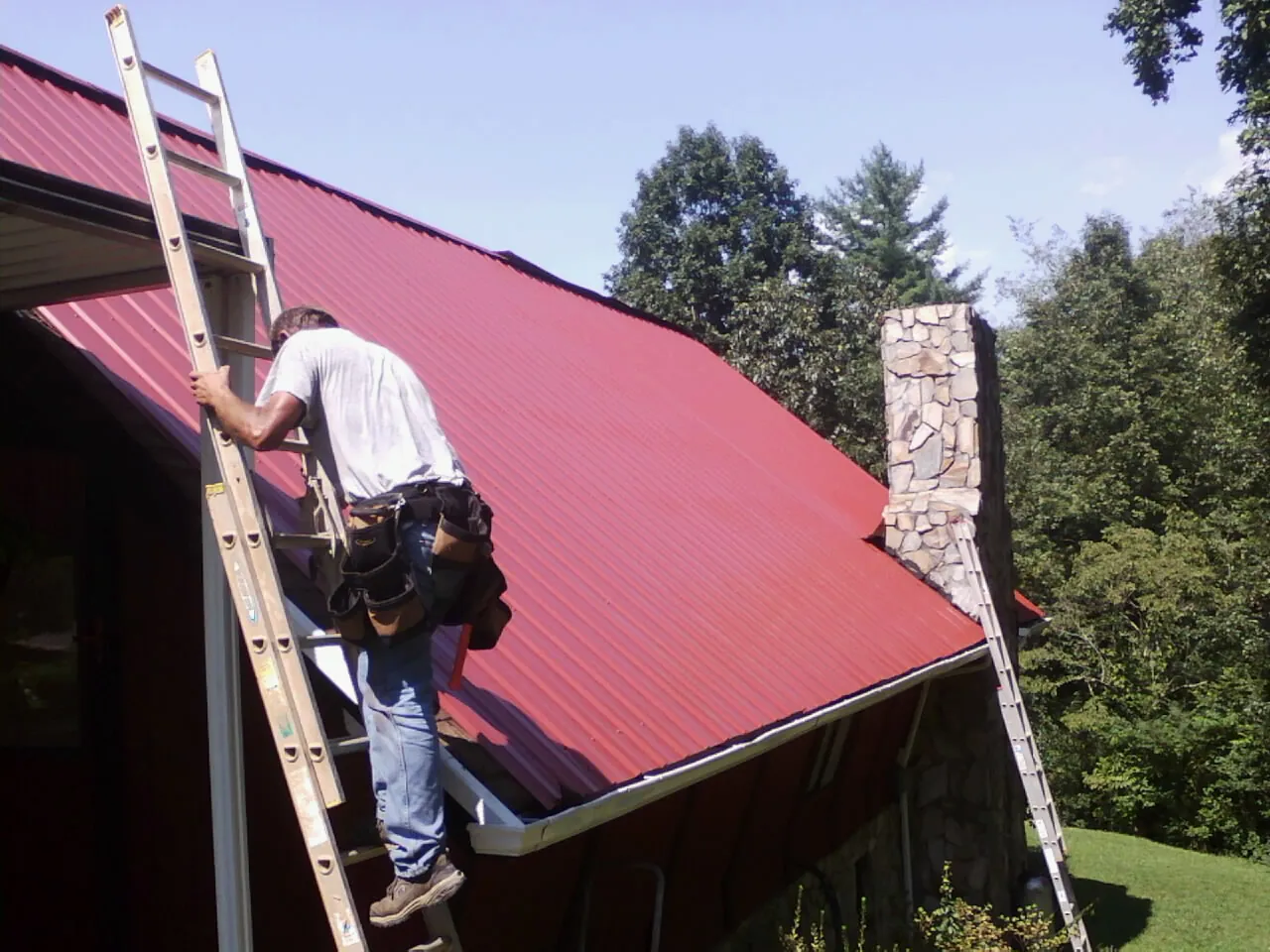 Roofer on a ladder beside a red metal roof and stone chimney — Soffit & Fascia Repair by Roofing Brothers in Kanarraville